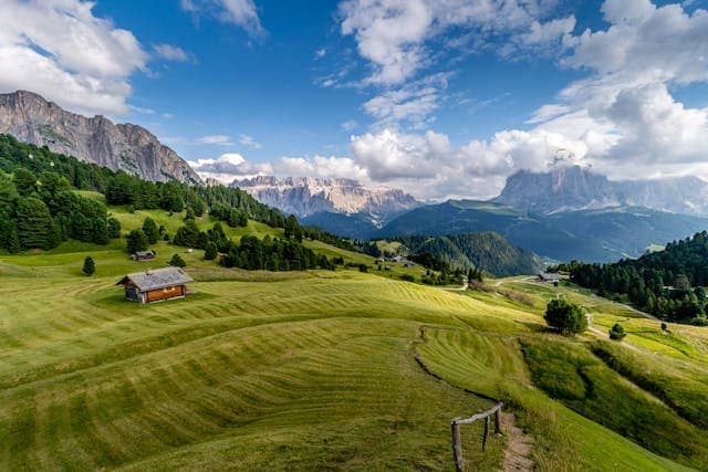 Paesaggio mozzafiato dell'Alto Adige con verdi prati ondulati, baite di montagna e le imponenti Dolomiti sullo sfondo. Un perfetto scenario per vivere Ferragosto in montagna, tra escursioni panoramiche, natura incontaminata e aria fresca.