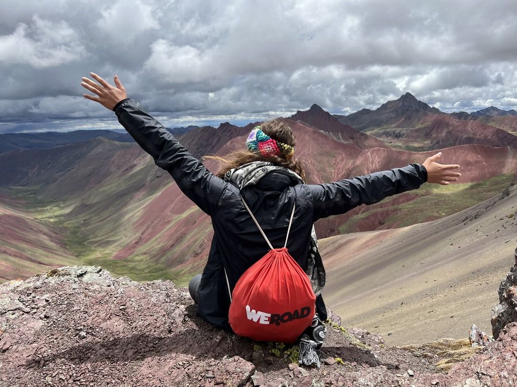 Viaggiatrice con zaino WeRoad osserva le montagne rosse del Perù dalla cima della Vinicunca, braccia aperte verso il paesaggio andino.