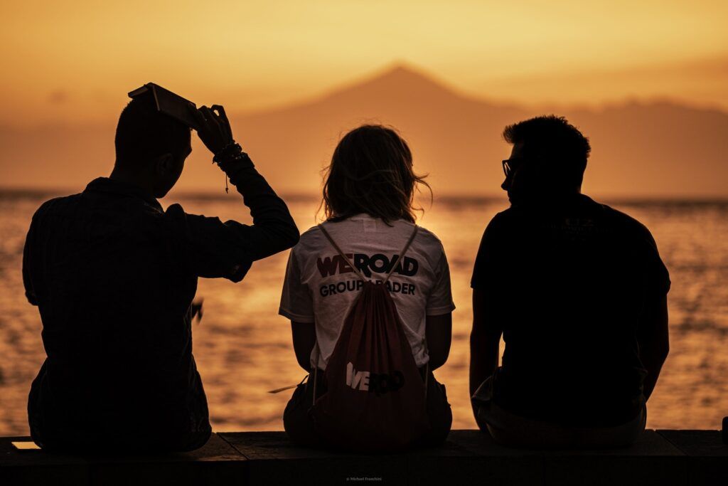 Tre viaggiatori WeRoad seduti di spalle davanti al mare al tramonto, con il profilo di un vulcano all’orizzonte