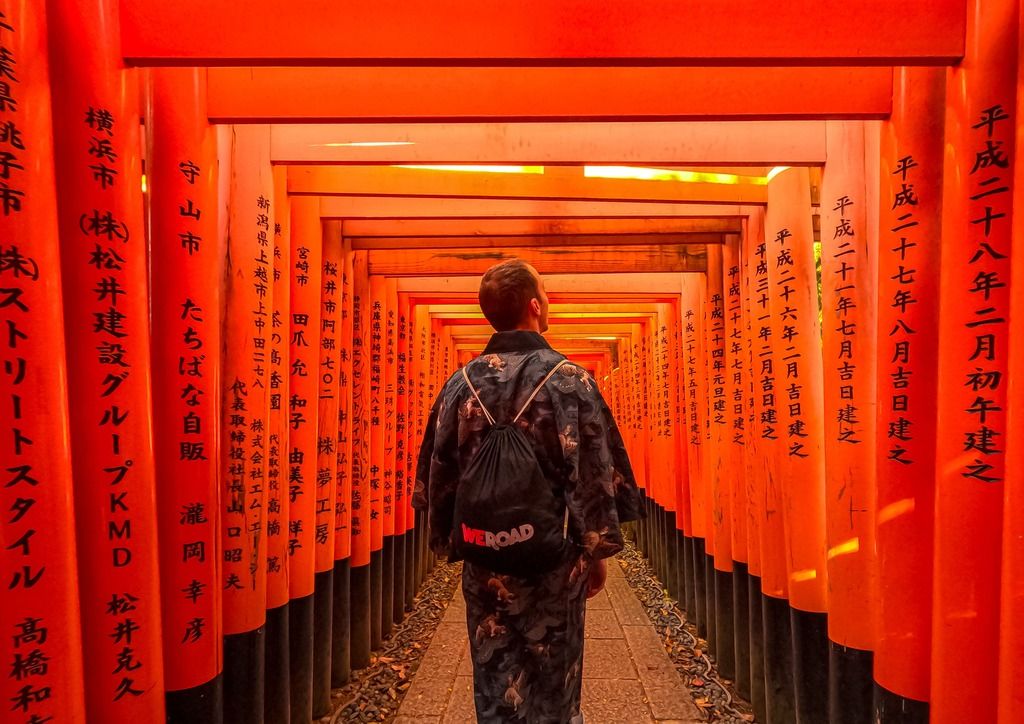 Viaggiatore con zaino WeRoad cammina tra i torii rossi del santuario Fushimi Inari a Kyoto, in Giappone.