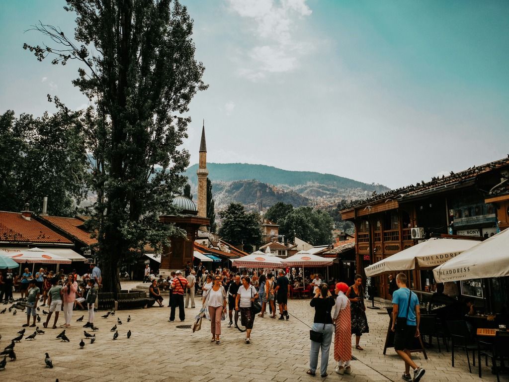 Strada di Sarajevo affollata da turisti e abitanti, tra bancarelle e una moschea sullo sfondo.