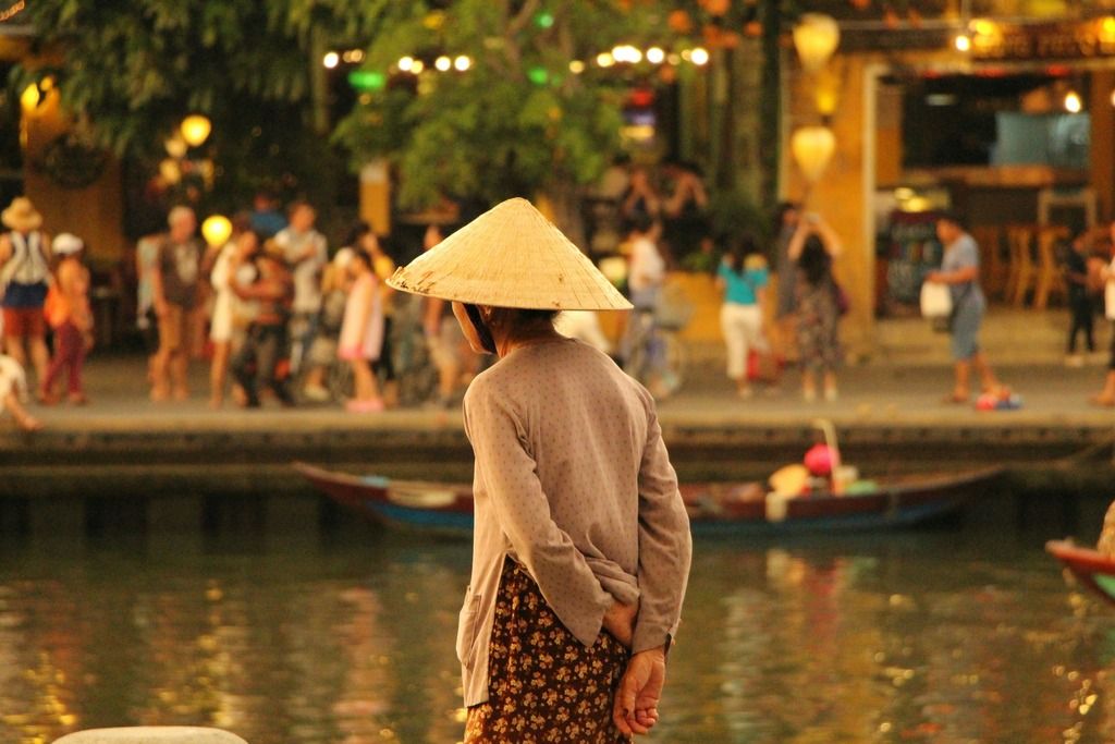 Persona con cappello tradizionale vietnamita osserva il fiume a Hoi An, con lanterne e passanti sullo sfondo.