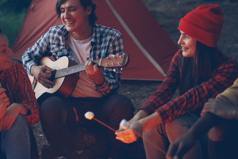 Gruppo di amici che ridono attorno a un fuoco serale vicino a una tenda da campeggio, uno suona la chitarra mentre gli altri arrostiscono marshmallow