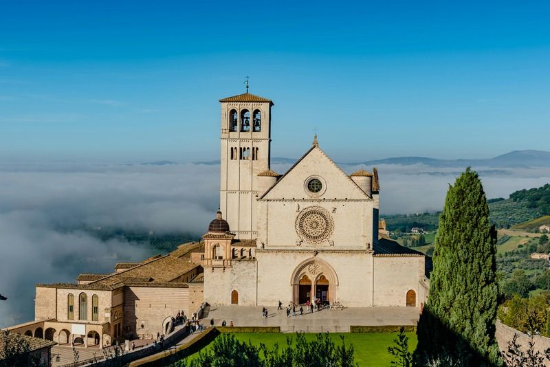 Basilica di San Francesco ad Assisi avvolta dalla nebbia nella valle umbra, con cielo azzurro e cipressi sullo sfondo
