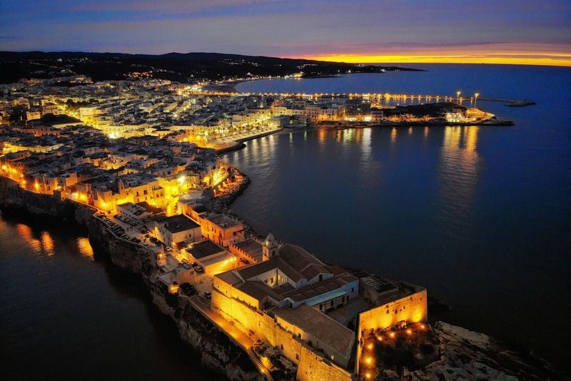 Vista aerea notturna di una città costiera pugliese con luci dorate riflesse sul mare Adriatico al tramonto