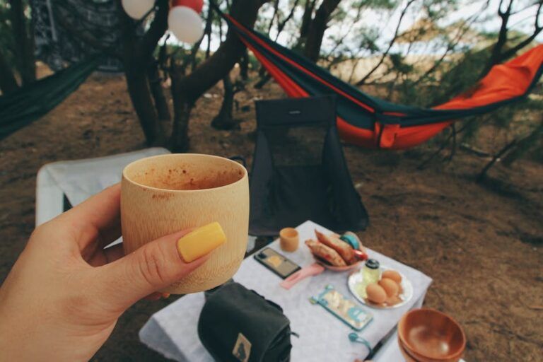 Mano che regge una tazza di bambù durante una colazione all'aperto, con amaca colorata e snack su un telo tra gli alberi