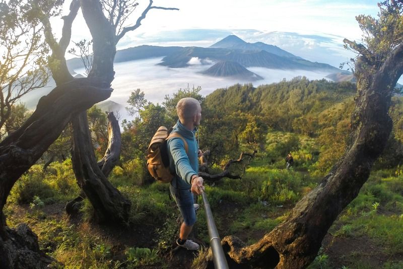 Escursionista con zaino e selfie stick sul versante del Monte Bromo in Indonesia, con nuvole nella valle sottostante
