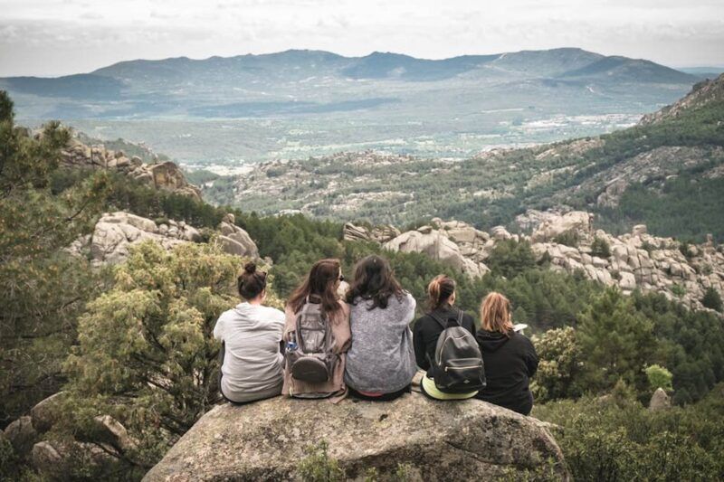 Cinque ragazze con zaini sedute su una roccia di spalle, che ammirano il panorama di una vallata montuosa con foreste e rocce