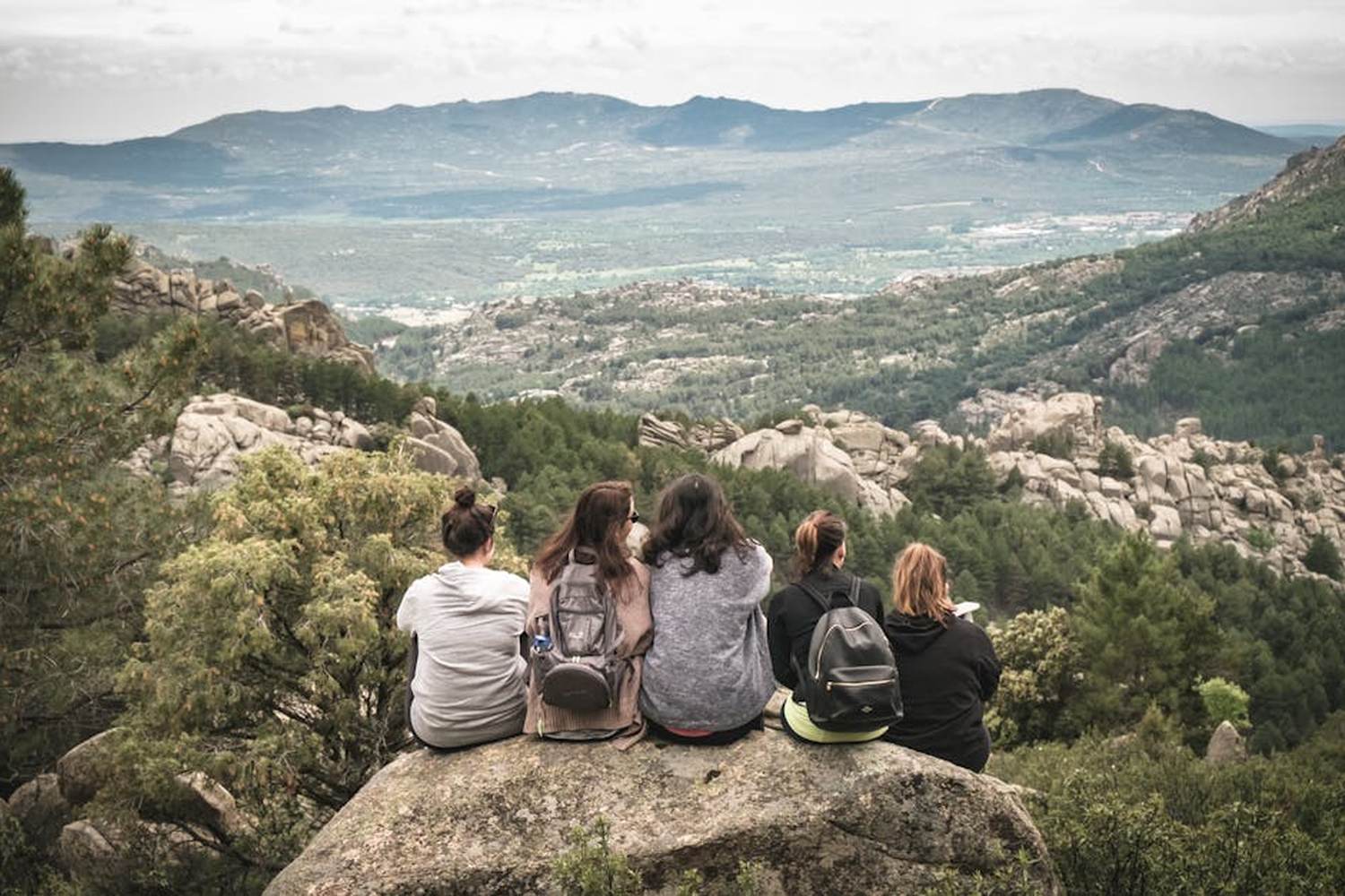 Cinque ragazze con zaini sedute su una roccia di spalle, che ammirano il panorama di una vallata montuosa con foreste e rocce