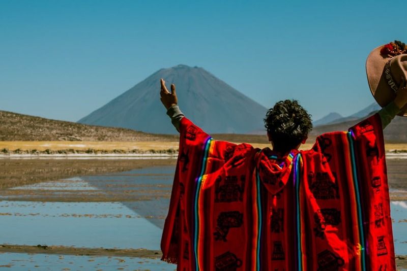 Viaggiatore con poncho andino rosso e cappello alzato di fronte a un vulcano e un lago