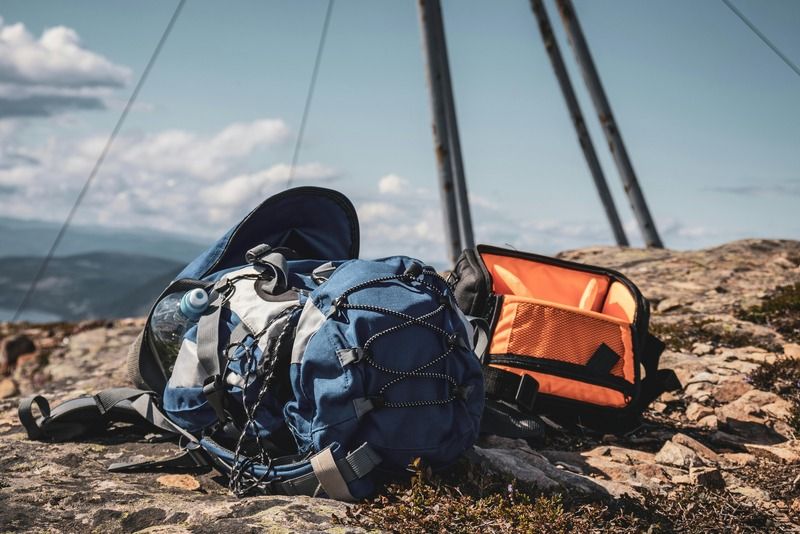 Zaino da trekking blu e borsa fotografica arancione posati su rocce in cima a una montagna con panorama sulle colline e cielo sereno