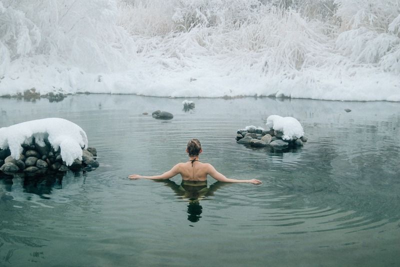 Donna di spalle con le braccia aperte in una piscina termale all'aperto circondata dalla neve in un paesaggio invernale innevato
