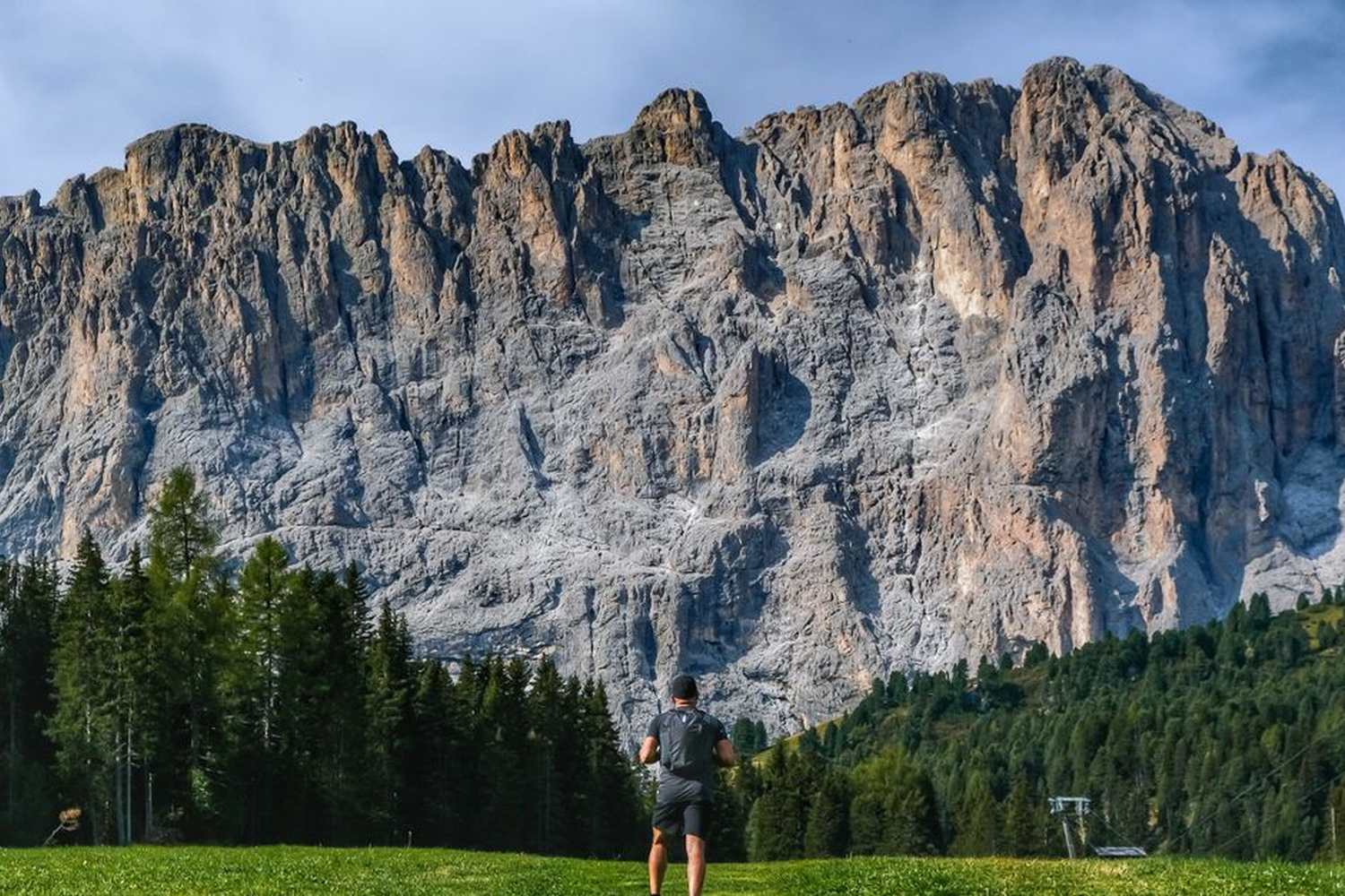 Escursionista in piedi su un prato verde di fronte alle maestose pareti rocciose delle Dolomiti, con foresta di abeti sullo sfondo