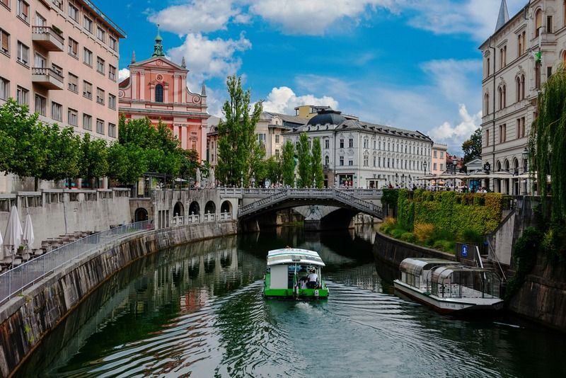 Barca elettrica verde sul fiume Ljubljanica a Lubiana sotto il ponte Tromostovje, con la Chiesa francescana rosa sullo sfondo
