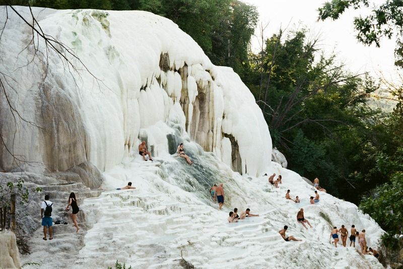 Gruppo di persone che fanno il bagno nelle sorgenti termali di travertino bianco di Bagni San Filippo, circondate da vegetazione lussureggiante in Toscana