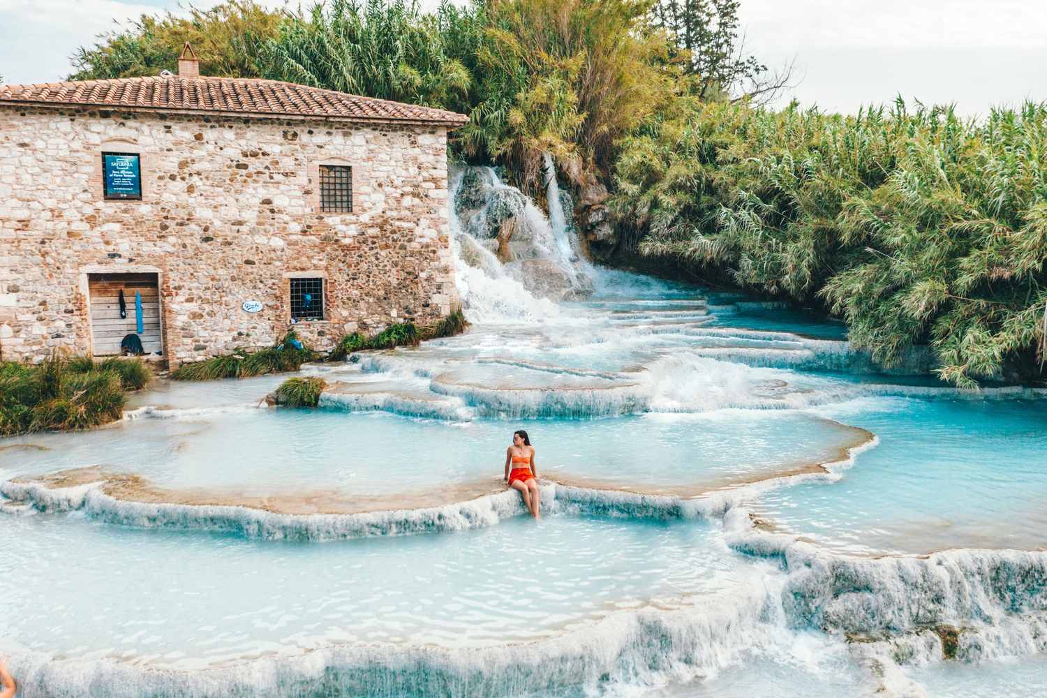 Donna seduta sulle vasche delle Terme di Saturnia con cascate di acque termali turchesi e antico casale in pietra sullo sfondo, Toscana