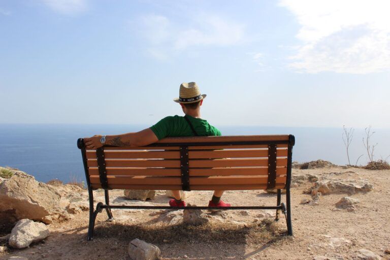 Viaggiatore con cappello di paglia seduto su una panchina di legno su un belvedere roccioso con vista sul mare azzurro