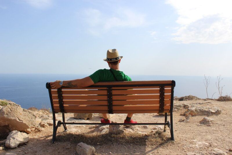 Viaggiatore con cappello di paglia seduto su una panchina di legno su un belvedere roccioso con vista sul mare azzurro
