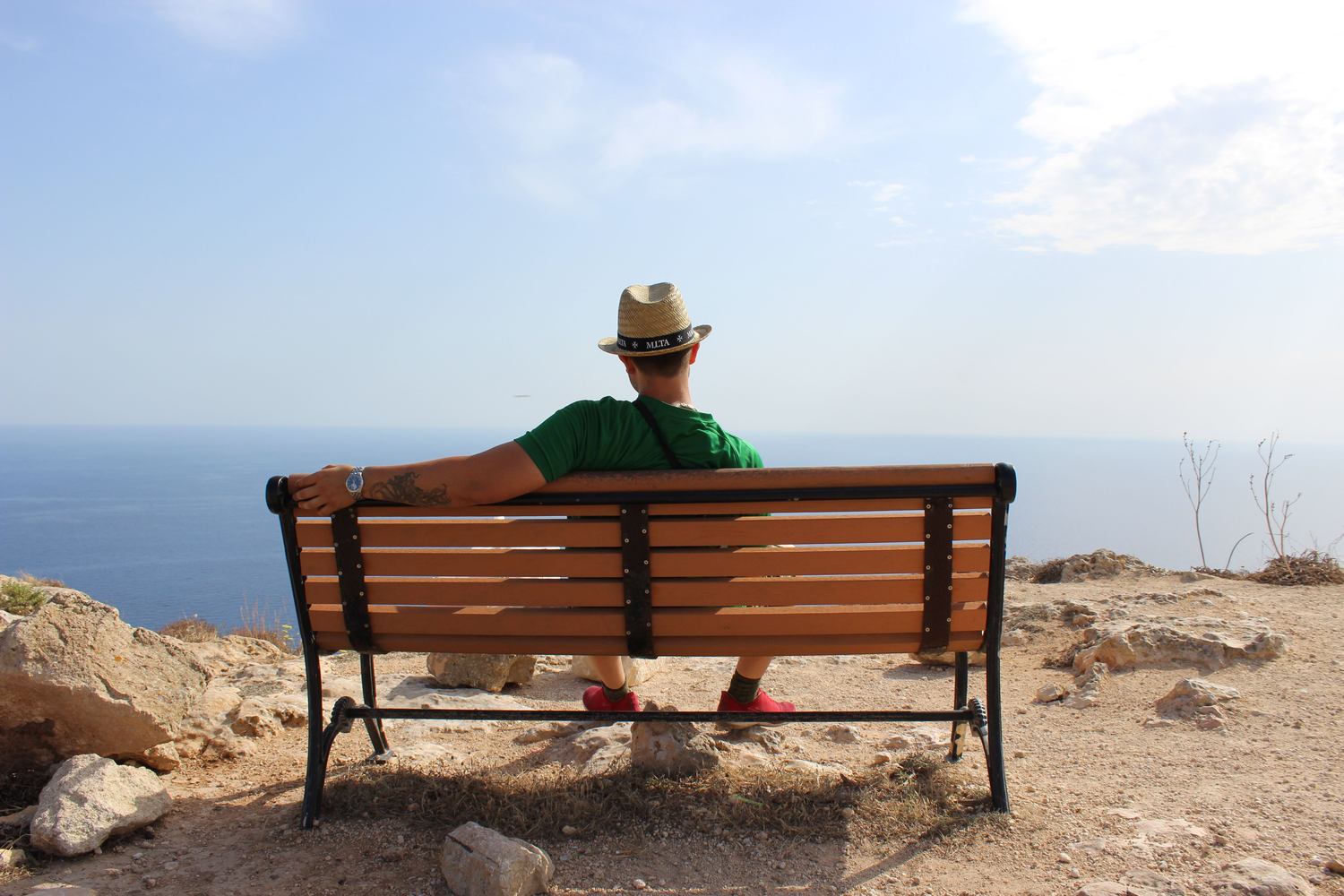 Viaggiatore con cappello di paglia seduto su una panchina di legno su un belvedere roccioso con vista sul mare azzurro