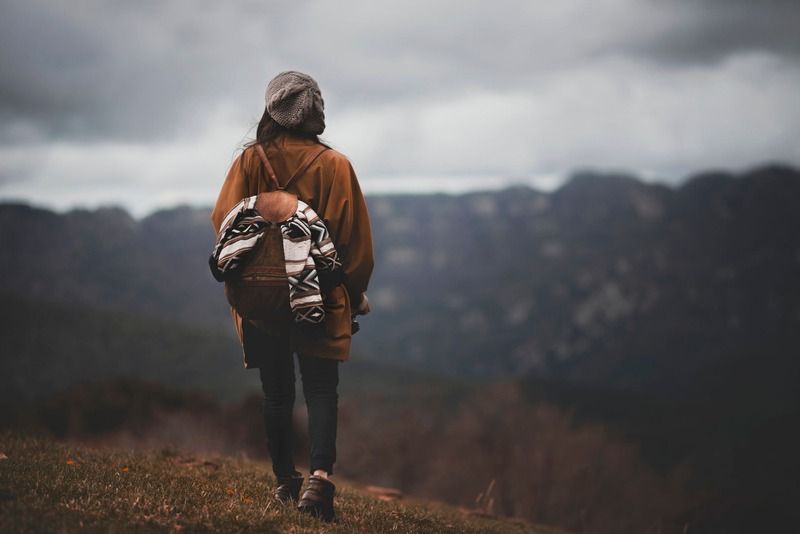 Viaggiatrice con zaino etnico e cappello di lana osserva un paesaggio montano avvolto da nuvole grigie