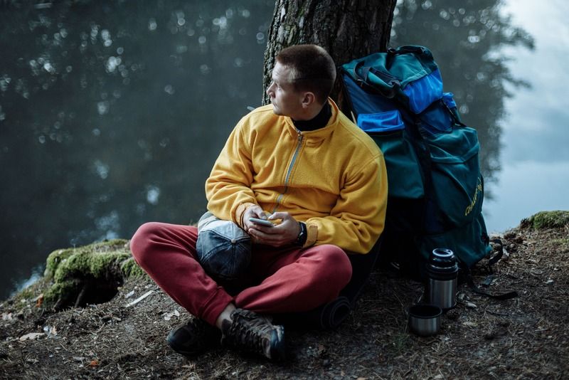 Escursionista in pausa seduto vicino a un lago con zaino e thermos durante il trekking