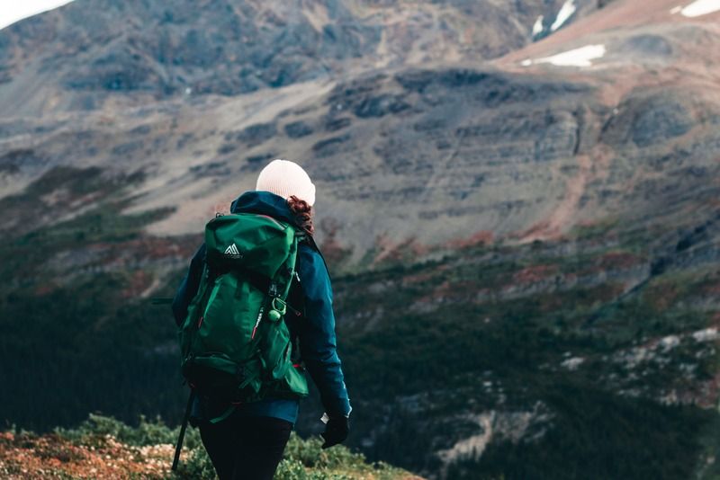 Escursionista con zaino verde cammina su un sentiero di montagna verso una vetta rocciosa