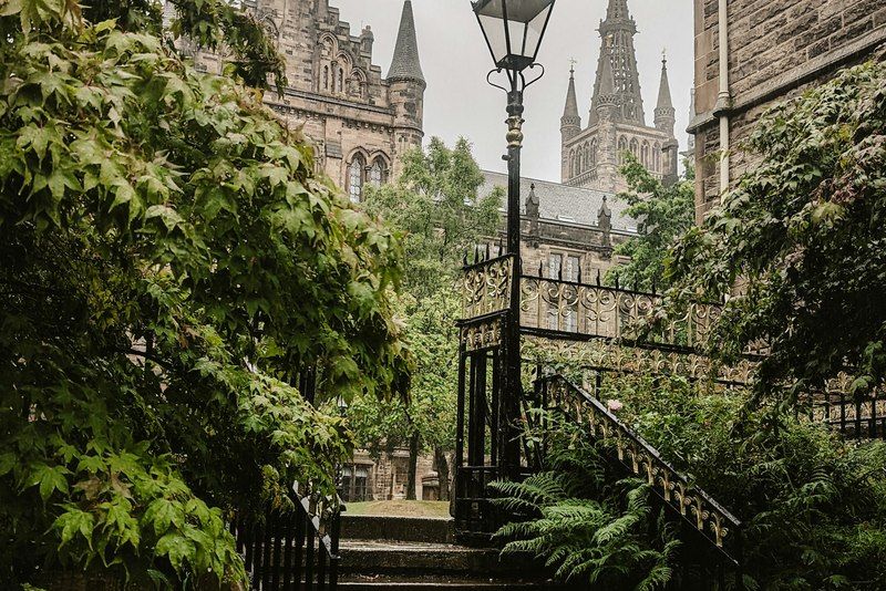 Scalinata di pietra tra felci e piante rigogliose, con un lampione d'epoca e le torri gotiche dell'Università di Glasgow sullo sfondo