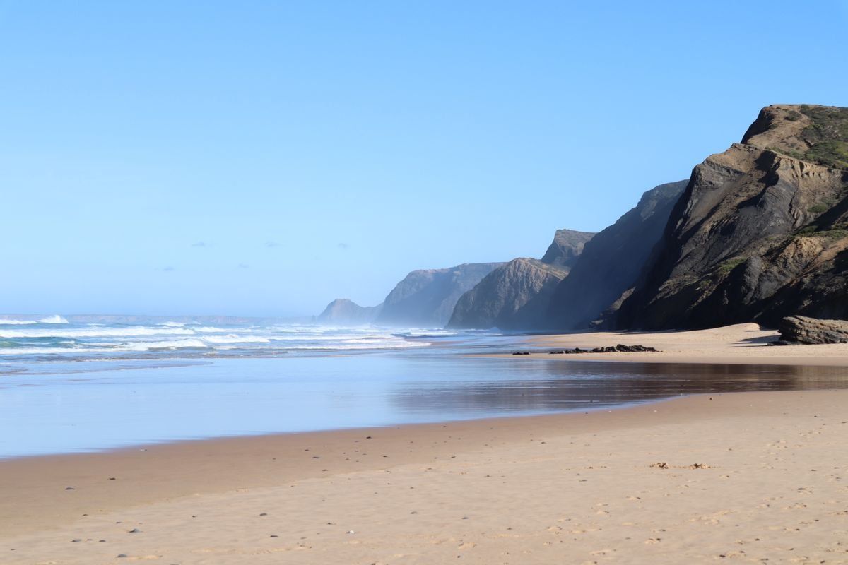 Ampia spiaggia deserta con scogliere scure e onde leggere sotto un cielo azzurro limpido