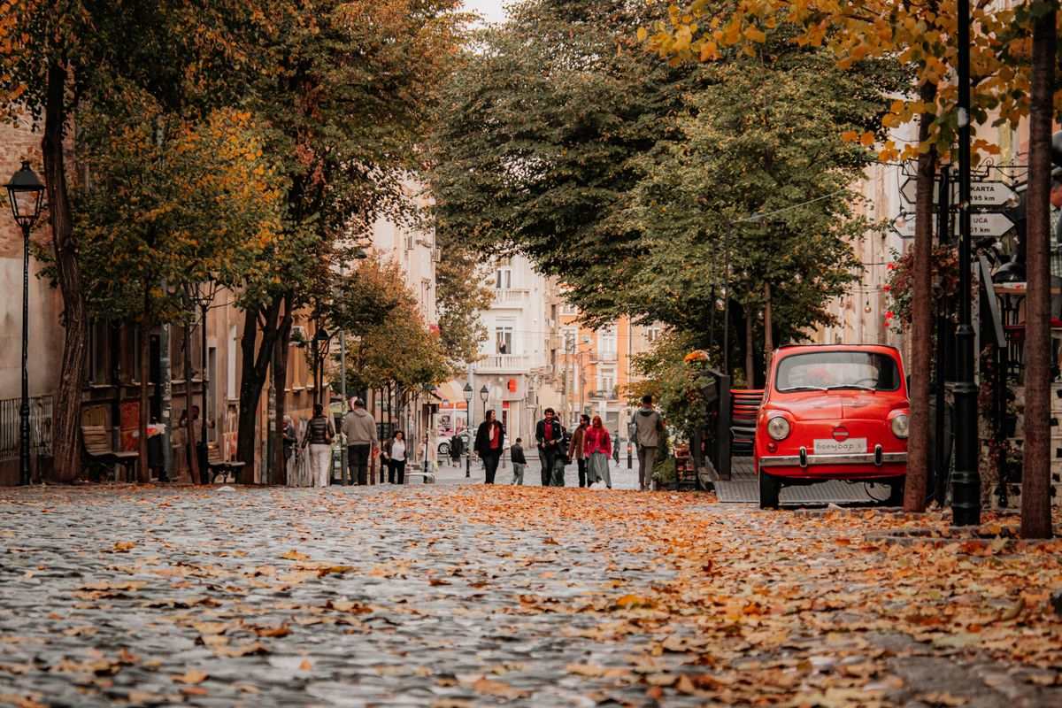 Auto rossa d'epoca su strada acciottolata con foglie cadute e passanti