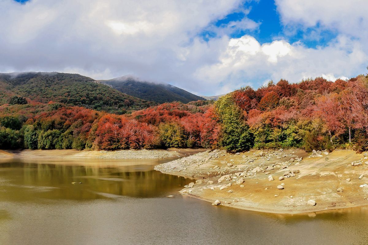 Lago con sponda rocciosa, alberi autunnali colorati e colline sotto cielo nuvoloso