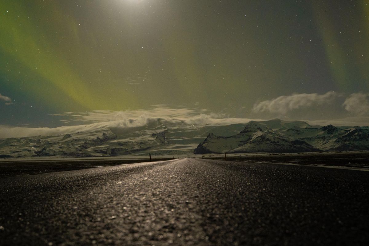 Strada notturna verso montagne innevate sotto una debole aurora boreale