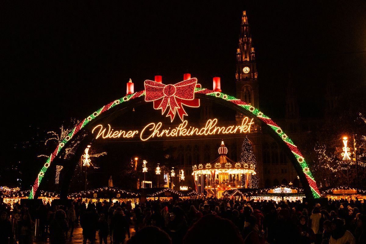 Arco luminoso del Wiener Christkindlmarkt con folla serale e torre illuminata sullo sfondo
