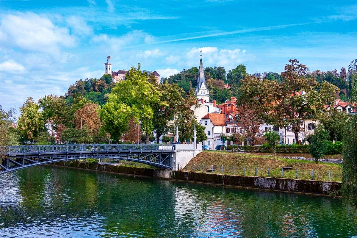 Fiume con passerella, case storiche e campanile ai piedi della collina alberata