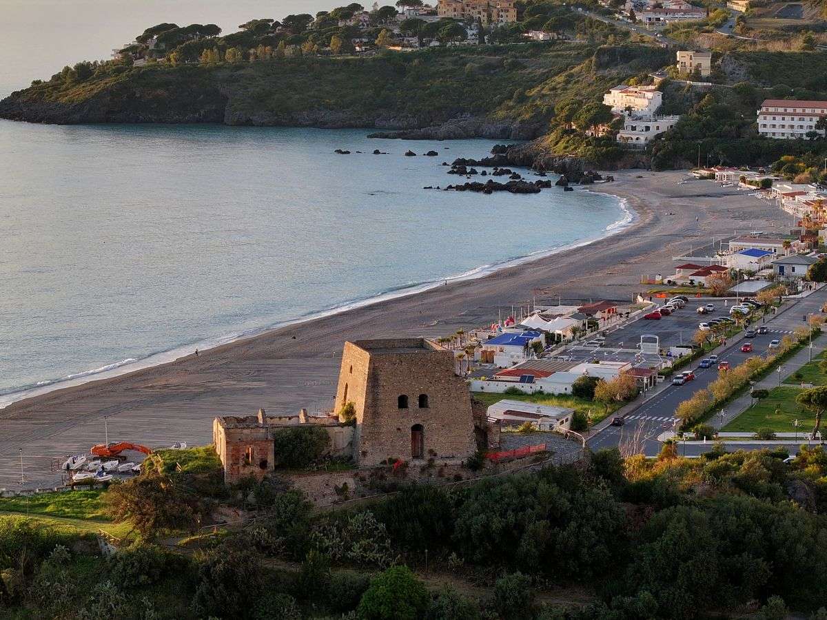 Spiaggia curva al tramonto con ruderi in primo piano e case sulla collina