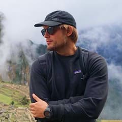Hombre con gorra y chaqueta oscura, con los brazos cruzados frente a crestas montañosas brumosas.