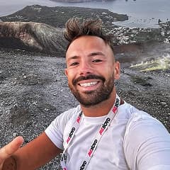 Man with a beard and mohawk wearing a WeRoad t-shirt takes a selfie with a volcanic island in the distance.