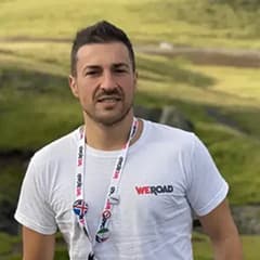 Uomo con t-shirt e laccetto WeRoad in piedi all'aperto, colline verdi sullo sfondo.