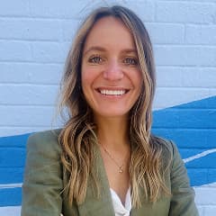Woman with long hair in an olive blazer smiles in front of a painted blue and white brick wall.