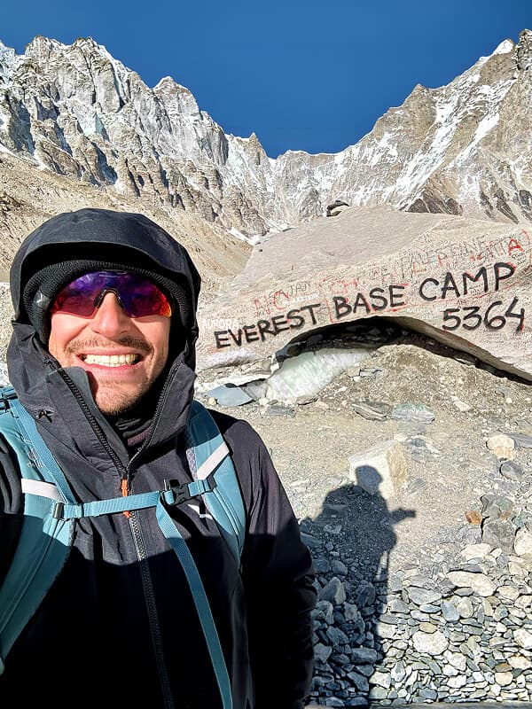 Uomo con giacca con cappuccio e occhiali da sole si fa un selfie davanti a vette innevate e a una roccia dipinta.