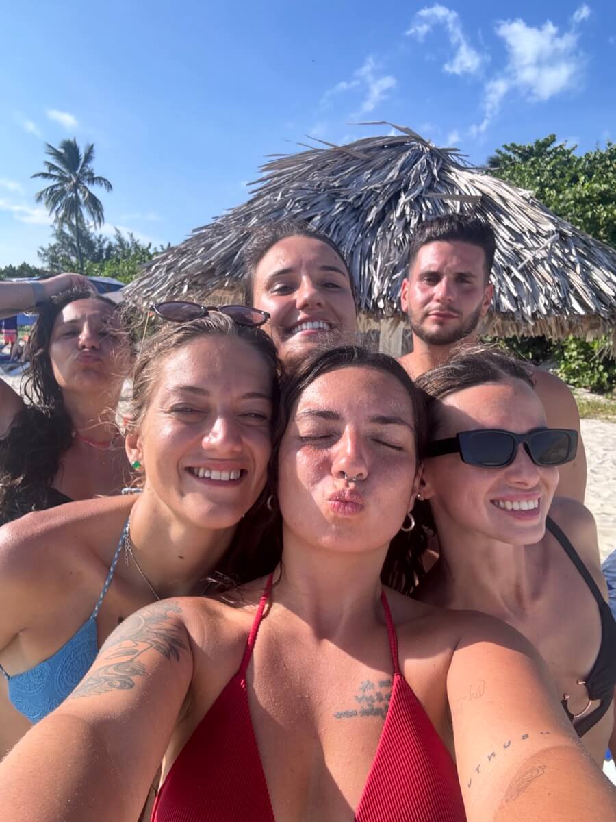 Selfie di un gruppo WeRoad: donne sorridenti sotto un ombrellone di paglia, con palme e cielo azzurro alle spalle.