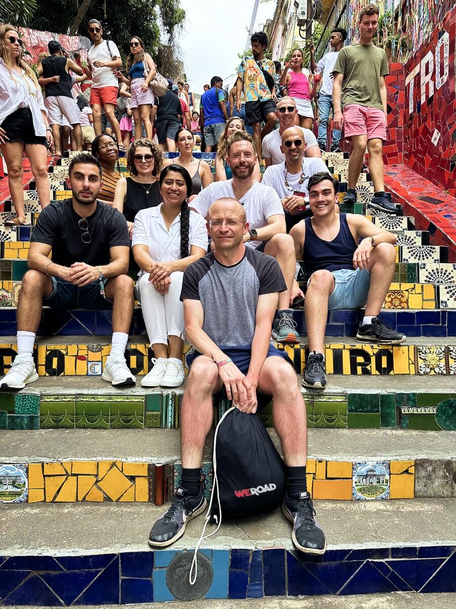 A WeRoad group trip seated on a colorful tiled mosaic staircase with a WeRoad-branded drawstring backpack in the foreground.