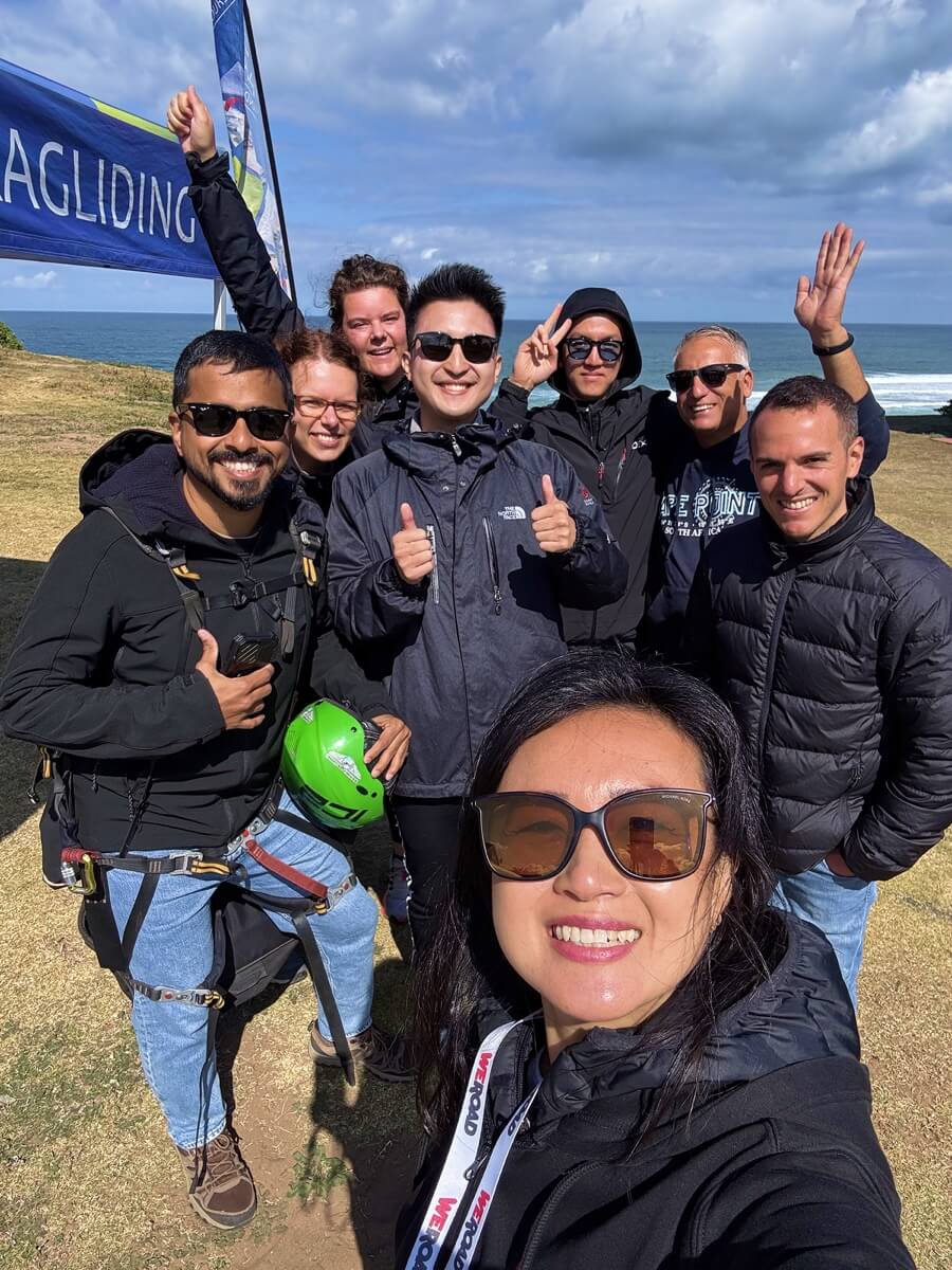A WeRoad group trip selfie of travelers smiling beside a paragliding banner on a coastal cliff.
