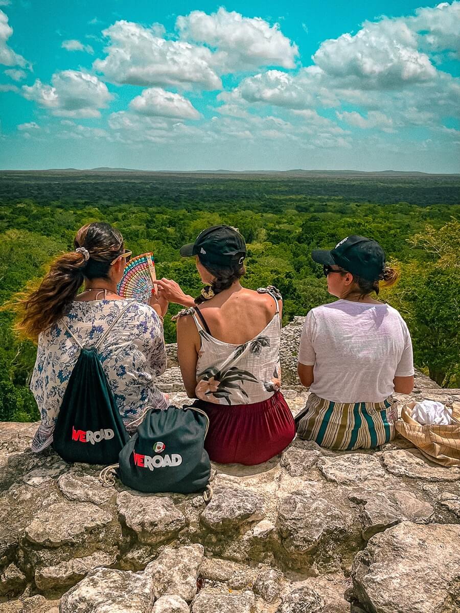 Trois femmes assises sur des ruines en pierre face à une vaste jungle verte, avec des sacs à dos WeRoad à côté.