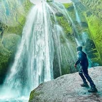 Person standing on a rocky ledge facing a tall waterfall cascading down a green moss-covered cliff.