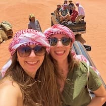 A WeRoad group trip selfie of two women in red checkered keffiyehs smiling on red desert sand with travelers in a jeep behind.
