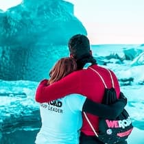 A WeRoad group leader and traveler embrace from behind facing turquoise icebergs in a glacier lagoon.