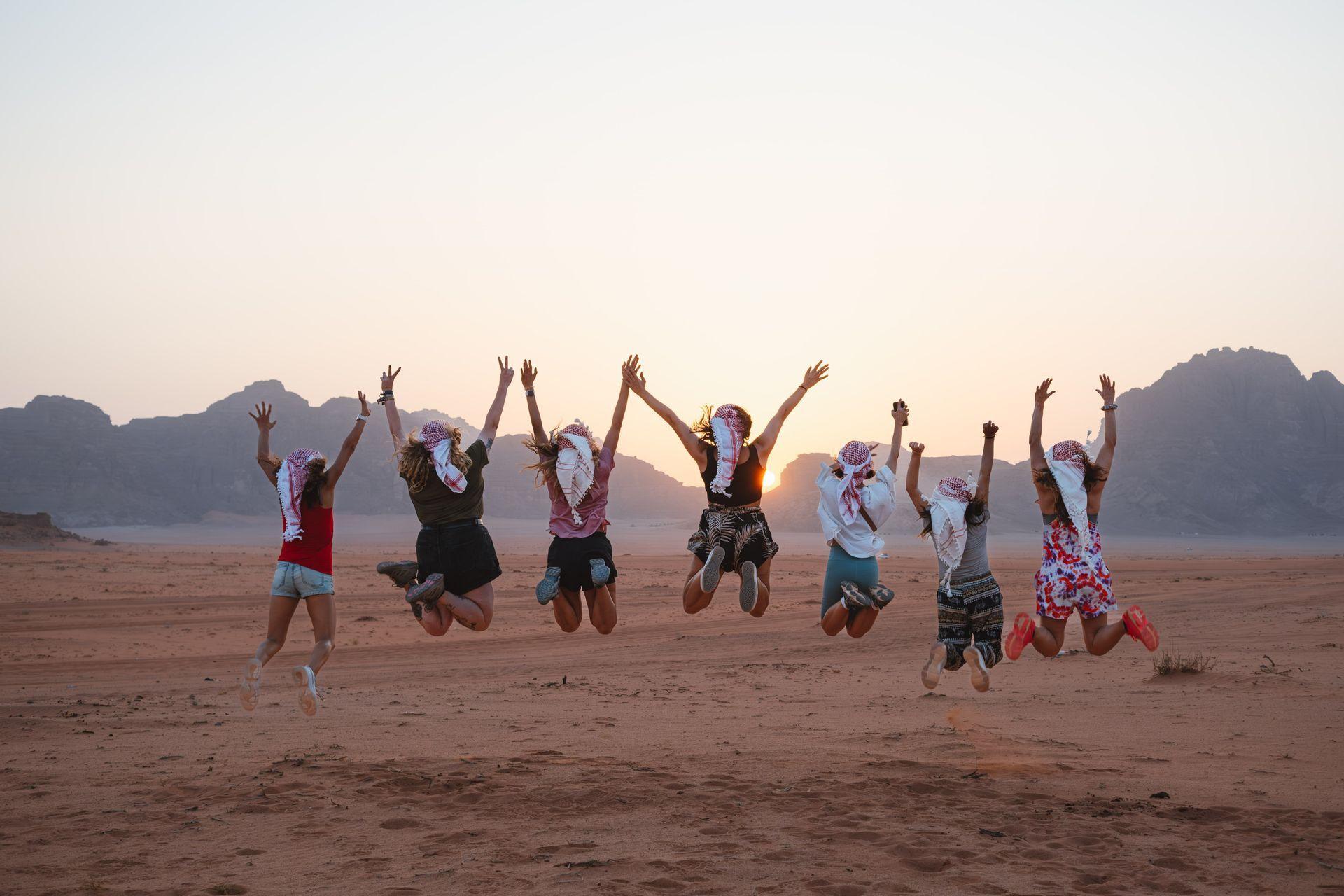A WeRoad group trip, seen from behind, jumps up in a desert with mountains in the background at sunset.