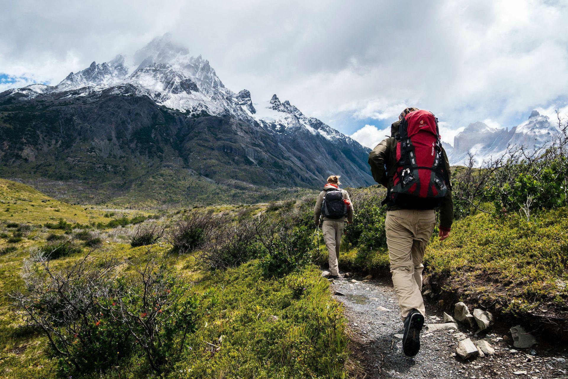 Deux randonneurs avec de grands sacs à dos marchent sur un sentier étroit à travers une vallée verdoyante vers des montagnes déchiquetées et enneigées.