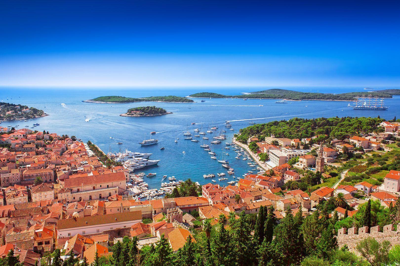 Veduta dall'alto di una cittadina costiera con tetti in terracotta, affacciata su un porto ricco di yacht e imbarcazioni, con isole all'orizzonte.