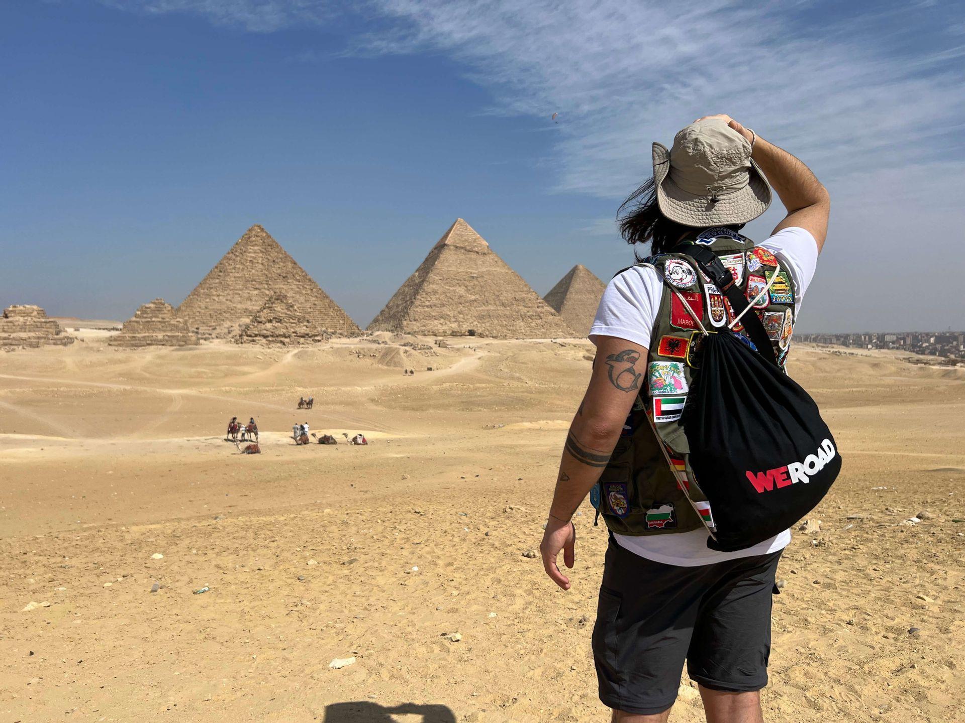 A traveler viewed from behind, wearing a hat and a WeRoad backpack, looks out at large pyramids in the desert.